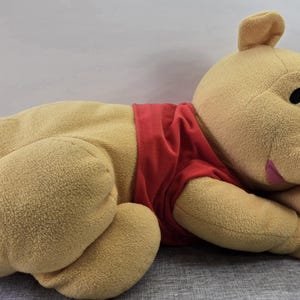May include: A plush Winnie the Pooh toy in a lying position. The bear is light yellow with a red shirt, black eyes, and a pink mouth. The toy is soft and cuddly, perfect for children.