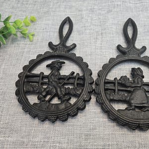 Puede incluir: Dos salvamanteles de hierro fundido negro con un diseño decorativo. Cada salvamanteles presenta un marco circular con un borde festoneado y un asa. Uno representa a un hombre con una azada, el otro a una mujer con flores.