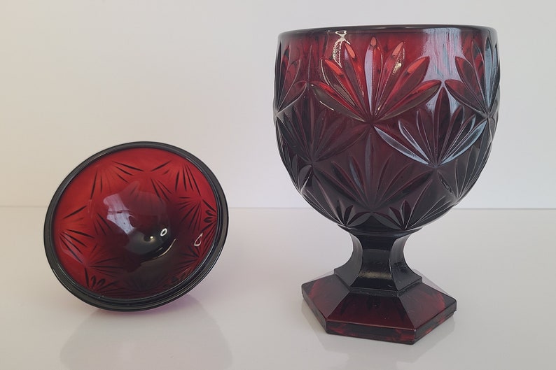 May include: A red glass candy dish with a footed base and a lid. The dish has a geometric pattern.
