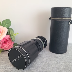 Puede incluir: Un objetivo de cámara negro con una tapa de objetivo negra que dice "SOLLIGOR 106" en letras blancas. El objetivo está al lado de una funda de cuero negra para objetivo de cámara con cremallera.