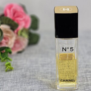 Puede incluir: Un frasco de perfume rectangular con tapa negra y detalles dorados. El frasco transparente revela un líquido amarillo. El texto "N°5" y "CHANEL PARIS" están impresos en la parte delantera. El fondo está borroso, con elementos florales rosas y verdes.