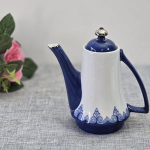 May include: A white and navy blue ceramic teapot with a silver-colored lid. The teapot has a curved spout and handle, with a decorative blue pattern around the base. The teapot is on a gray surface.