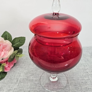 Può includere: Un vaso da farmacia in vetro rosso rubino con coperchio e stelo in vetro trasparente. Il vaso ha un corpo arrotondato e una maniglia in vetro trasparente sul coperchio. Il vaso è appoggiato su una superficie grigia.