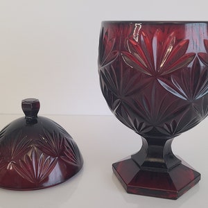 May include: A red glass candy dish with a lid. The dish has a pedestal base and a geometric pattern.