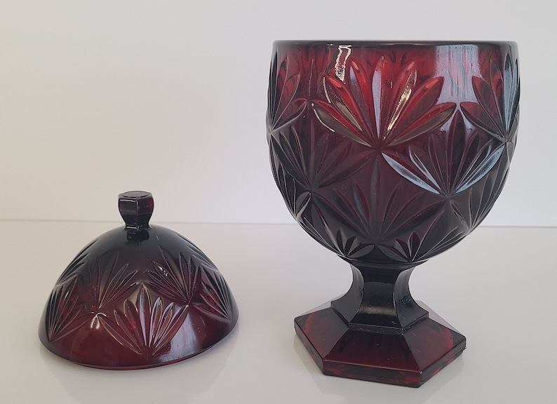 May include: A red glass candy dish with a lid. The dish has a pedestal base and a geometric pattern.