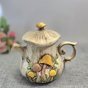 Può includere: Una teiera in ceramica a forma di fungo con corpo color crema e superficie testurizzata. Il coperchio presenta un piccolo pomello a forma di cappello di fungo. La parte anteriore è decorata con figure di funghi in rilievo gialle e marroni.