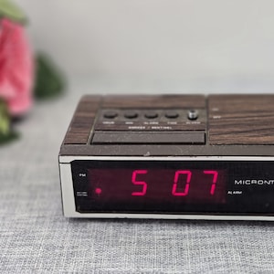 May include: A vintage Micronta digital alarm clock with a wood-grain finish. The red LED display shows the time as 5:07. The clock has several buttons and a black power cord.
