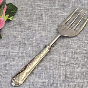 Puede incluir: Un tenedor de metal plateado con un mango largo y delgado que presenta un patrón geométrico en relieve compuesto por formas de diamante y motivos florales. La cabeza del tenedor tiene cuatro puntas y un acabado pulido.