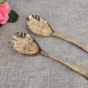 Puede incluir: Dos utensilios antiguos de plata para servir, una cuchara y un tenedor, con diseños florales ornamentados. Los utensilios están colocados sobre una superficie de lino gris, con flores rosas en el fondo. La cuchara tiene un cuenco ancho y el tenedor tiene tres púas.