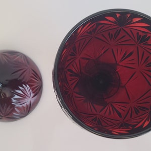 May include: A set of two red glass candle holders with a geometric pattern. The candle holders are made of clear glass with a red tint and have a starburst pattern.