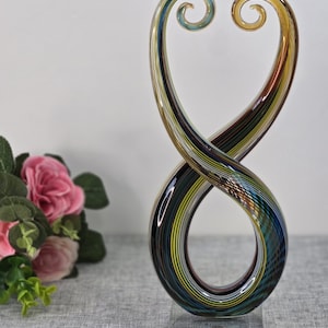 Può includere: Scultura decorativa in vetro a forma di simbolo dell'infinito. La scultura presenta fasce a spirale di vetro blu, giallo, marrone e verde. Poggia su una base rettangolare trasparente. Un'opera d'arte moderna.