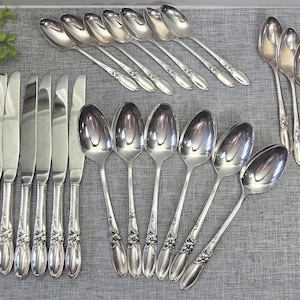 May include: A collection of silver-toned cutlery including six dinner knives with smooth blades and six dinner spoons, plus seven smaller spoons and seven additional spoons, all featuring a floral embossed pattern on the handles.