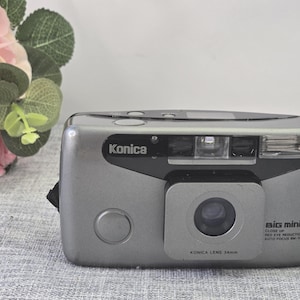 Op de afbeelding: Een zilveren Konica Big Mini SR 35mm filmcamera. De camera heeft een zwarte bovenkant en een grote lens. De tekst "Konica" staat erop. De camera heeft een 34mm lens en beschikt over close-up en rode-ogenreductie.