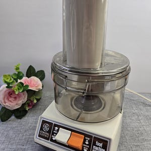 Peut inclure: Un robot culinaire General Electric vintage, avec une base blanche et un bol en plastique transparent. Il comporte un grand récipient gris cylindrique sur le dessus. Le panneau de commande comporte des boutons blancs et orange.