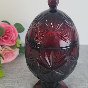 Pode incluir: Um prato de doces de vidro vermelho escuro com tampa. O prato tem um padrão geométrico e fica em uma base de pedestal.