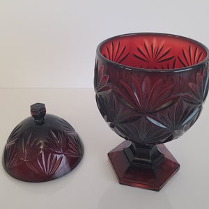 May include: A red glass candy dish with a lid. The dish has a cut glass design with a starburst pattern. The dish is on a white background.