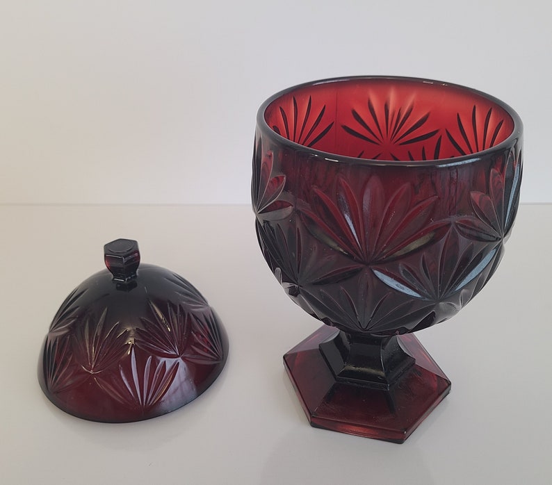 May include: A red glass candy dish with a lid. The dish has a cut glass design with a starburst pattern. The dish is on a white background.