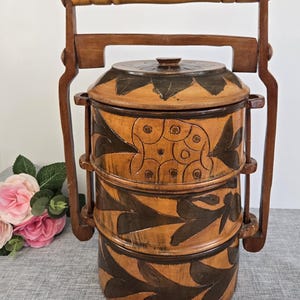 Könnte beinhalten: Eine dreistöckige Holz-Lunchbox mit einem geschnitzten Blumenmuster. Die Box hat einen braunen Holzgriff und ist aus Naturholz gefertigt.