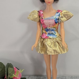 May include: A vintage doll in a gold dress with a floral bodice and puffed sleeves. The doll has short brown hair and blue eyes. The dress has a gold skirt and a bodice with pink, blue, and white floral patterns.