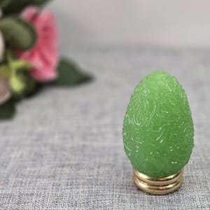 以下が含まれることがあります： 表面全体に複雑な花のレリーフ模様が施された、淡い緑色の卵型の装飾品。この装飾品は、安定した土台となる円形のゴールドトーンの金属製ベースに取り付けられています。