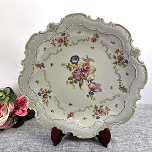 May include: A round porcelain plate with a scalloped, embossed edge featuring a central floral bouquet of pink, yellow, and blue flowers. The rim is decorated with a subtle lattice pattern and gold-toned accents, surrounding smaller floral sprigs.