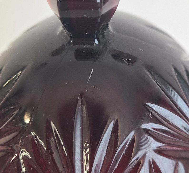 May include: A dark red glass cloche with a faceted design. The cloche has a knob on top.