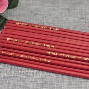 Peut inclure: Une collection de crayons en bois rouge avec des inscriptions dorées. Chaque crayon porte le texte PATRIA VENUS PENCIL COMPANY LTD. MADE IN CANADA 6351-HB. Les crayons sont disposés en pile parallèle, mettant en valeur leur couleur uniforme.