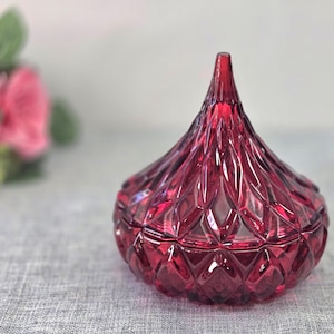 May include: A deep red, teardrop-shaped glass jar with a faceted, diamond-cut pattern across its surface. The container features a pointed lid that sits flush with the base, creating a uniform geometric texture throughout the entire object.