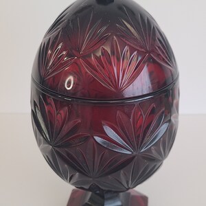 May include: A red glass candy dish with a lid. The dish is decorated with a cut glass pattern and has a pedestal base.