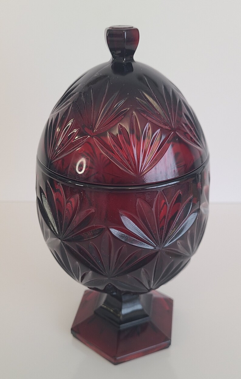 May include: A red glass candy dish with a lid. The dish is decorated with a cut glass pattern and has a pedestal base.
