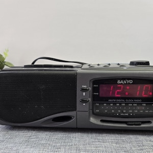 Puede incluir: Una radio reloj digital AM/FM Sanyo gris que muestra las 12:10 PM en dígitos rojos. La radio tiene una rejilla de altavoz negra, botones de control y un dial de volumen. El fondo es una superficie gris claro.