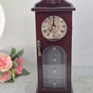 May include: A dark brown wooden mantel clock with a rounded top featuring a shell motif. The clock face has a silver background with black Roman numerals and ornate scrollwork. A glass door below displays three small silver circular accents.