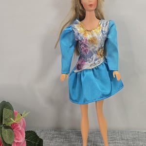 Puede incluir: Una muñeca vintage con cabello rubio y ojos azules, vestida con un vestido azul con mangas largas y un corpiño floral. El vestido de la muñeca tiene una falda plisada. La muñeca está de pie.
