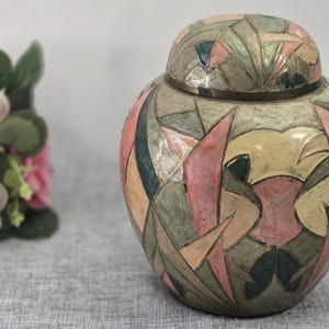 Puede incluir: Una urna decorativa de metal con tapa extraíble, que presenta un patrón de mosaico geométrico en tonos apagados de rosa, verde salvia, crema y verde azulado oscuro. La superficie tiene un aspecto texturizado con finas líneas metálicas.
