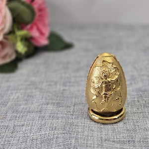 May include: A gold-colored egg-shaped ornament featuring a raised relief design of a winged figure. The object has a textured surface and rests on a smooth, circular gold-colored base. The finish is metallic and reflective throughout the piece.