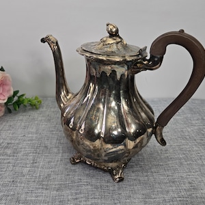 Peut inclure: Une théière ancienne en argent avec une poignée en bois. La théière a un corps cannelé, un bec verseur incurvé et un couvercle avec un bouton décoratif. La théière est posée sur une surface grise.
