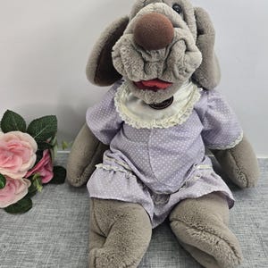 May include: A plush, gray dog puppet with a large brown nose and floppy ears. The puppet wears a lavender dress with white polka dots and a white lace collar. The dog's mouth is open, revealing a red tongue.