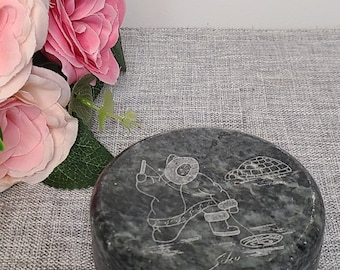 Può includere: Una scultura rotonda in steatite verde che rappresenta una persona che pesca sul ghiaccio. La scultura è dettagliata con una persona che tiene una canna da pesca, un igloo e un buco nel ghiaccio. La scultura è firmata "Sikuu" sul fondo.