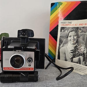 May include: A vintage Polaroid Colorpack 80 Land Camera with a black and silver body, flash attachment, and strap. The camera is next to a box with a rainbow design and a booklet titled "Meet the Colorpack 82 and 80."