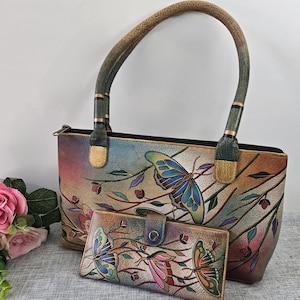 May include: A leather handbag and matching wallet set. The set features a colorful butterfly and floral design. The handbag has a curved handle and a zipper closure. The wallet has a snap closure.