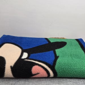 May include: A folded blue, green, and black fleece blanket featuring a cartoon character's face. The blanket has a blue border and a green section. The character's face is in the centre, with black, white, and pink details.