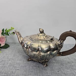 May include: A silver-colored teapot with a brown handle and a decorative lid. The teapot has a rounded body with a fluted design and sits on three small feet. A bouquet of pink roses is in the background.