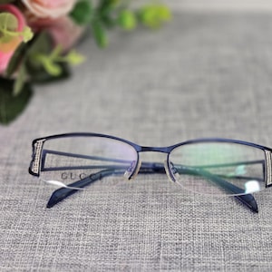 May include: Navy blue eyeglasses with a rectangular frame and clear lenses. The brand name "GUCCI" is visible on the frame. The temples feature a decorative design with small, sparkling accents. The glasses are set against a grey background.