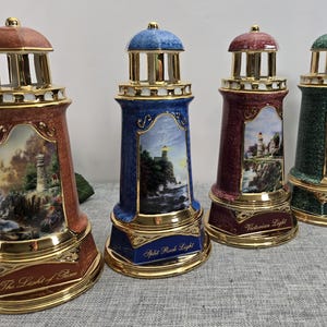 Peut inclure: Quatre figurines décoratives en forme de phare, de couleurs rouge, bleu, bordeaux et vert, avec des accents dorés. Chaque phare présente une peinture de paysage détaillée et du texte à la base. Les phares mesurent environ 20 cm de haut.