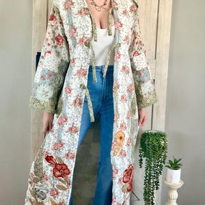 May include: A long, floral-patterned coat with a patchwork design. The coat features a white background with red and pink roses, green accents, and a light blue floral pattern. It has a hood and is open in the front, with tie closures. The sleeves are three-quarter length.