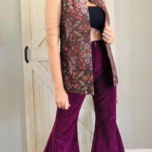 May include: A sleeveless vest featuring a dark brown, red, and grey paisley pattern, paired with deep purple corduroy bell-bottom pants and a black crop top. The vest has a straight cut with two front pockets and a button closure at the waist.
