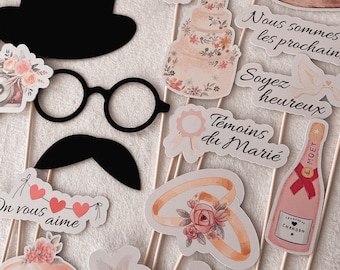 May include: A set of wedding photo booth props featuring a variety of designs, including a top hat, glasses, a mustache, a bouquet of flowers, a wedding cake, a champagne bottle, rings, and signs that say "Nous sommes les prochains", "Soyez heureux", "Témoins du Marié", and "Vive les Mariés!"
