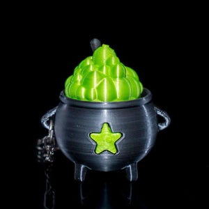 May include: A small, silver cauldron with a green star on the front. The cauldron is filled with a green, bubbling liquid. The cauldron is on a black background.