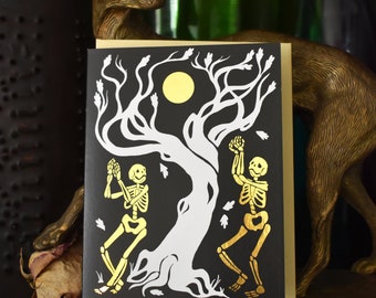 May include: A black card with a gold foil design of two skeletons dancing under a tree with a full moon. The tree is white and the background is black.