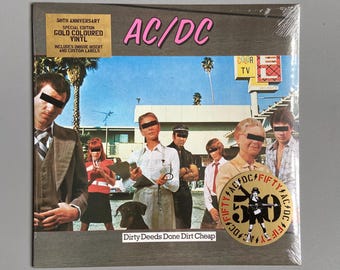 Puede incluir: Una portada de álbum de vinilo dorado para el álbum de AC/DC "Dirty Deeds Done Dirt Cheap". La portada presenta una foto de la banda frente a un motel con un letrero de "Color TV". El álbum es una edición especial del 50 aniversario.